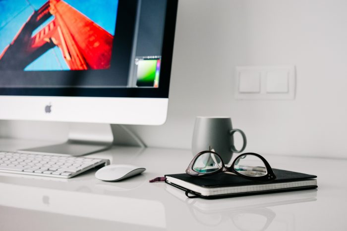 A photo of glasses placed on a journal along with a Mug and an iMac